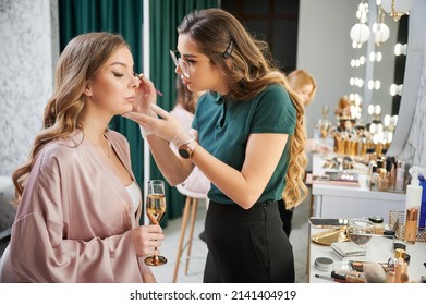 Beauty specialist applying eyeshadow on client eyelid in visage studio. Young woman sitting at dressing table and holding glass of champagne while makeup artist doing professional makeup.