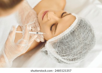 Beauty specialist injects neurotoxin or dermal filler in crows feet or upper eyelid. Close up woman's head in white cap and doctor's hands in gloves. Aesthetic face skin eye wrinkle treatment concept - Powered by Shutterstock