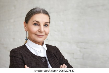 Beauty, Style, Fashion And Age Concept. Close Up Portrait Of Positive Elegant 60 Year Old Female With Gray Hair And Wrinkled Face Posing Against White Brick Wall Background And Smiling At Camera