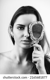 Beauty, style and make-up concept. Black and white portrait of beautiful woman covers her one eye with old vintage dirty mirror. Model with natural make-up, long dark hair and looking to camera