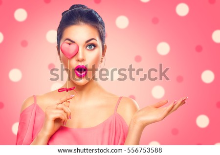 Beauty surprised Young fashion model Girl with Valentine Heart shaped cookie in hand. Love. Beautiful young woman pointing hand, advertising gesture. Valentines Day gift. Pink polka dots background.