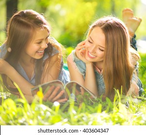 Beauty Teen Girls Reading Magazine And Having Fun Outdoors. Beautiful Joyful Teenagers Lying On Spring Grass, Laughing And Reading Fashion Magazine In Spring Park. Girlfriends Outdoor. Friendship