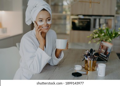 Beauty treatment personal care and hygiene concept. Lovely young woman applies cosmetic patches under eyes wears bathrobe drinks coffee talks to friend via smartphone sits at table over home interior - Powered by Shutterstock