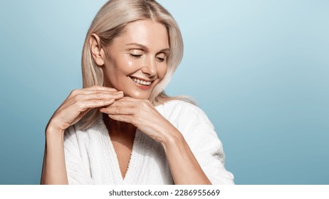 Beauty and wellbeing. Mature woman with glowing, clear skin without aging wrinkles, no blemishes, smiling coquettish, wearing spa bathrobe, doing makeup skincare procedures at home, blue background. - Powered by Shutterstock