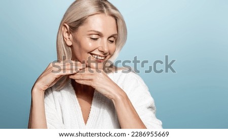 Beauty and wellbeing. Mature woman with glowing, clear skin without aging wrinkles, no blemishes, smiling coquettish, wearing spa bathrobe, doing makeup skincare procedures at home, blue background.