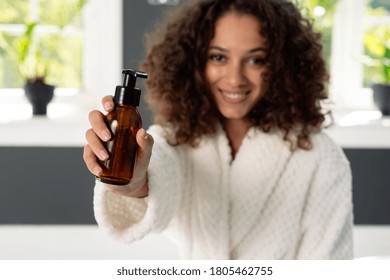 Beauty And Wellness Concept. Selective Focus On Bottle With Dispenser In Afro American Woman Hands. Happy African Girl Standing In Bathrobe At Bathroom, Holding Natural Skincare Or Body Care Cosmetics