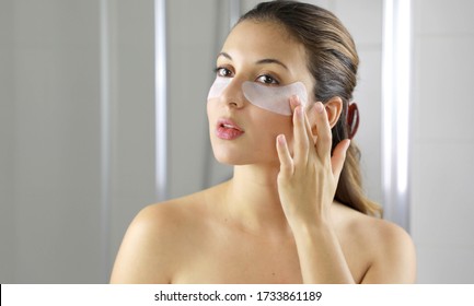 Beauty woman applying anti-aging under-eye mask looking herself in the mirror in the bathroom. Skin care girl touch patches of fabric mask under eyes to reduce eye bags.