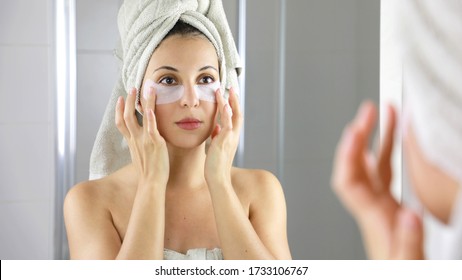 Beauty Woman Applying Anti-fatigue Under-eye Mask Looking Herself In The Mirror In Bathroom. Skin Care Girl Touch Patches Of Fabric Mask Under Eyes To Reduce Eye Bags.
