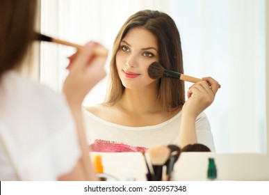 Beauty woman applying makeup. Beautiful girl looking in the mirror and applying cosmetic with a big brush. Girl gets blush on the cheekbones. Powder, rouge 