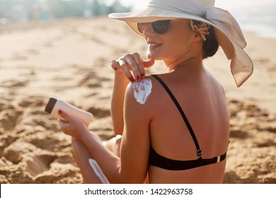 Beauty Woman Applying Sunscreen Creme On Tanned Shoulder. Skincare. Body Sun Protection Suncream. Bikini Hat Woman Applying Moisturizing Sunscreen Lotion On Back.