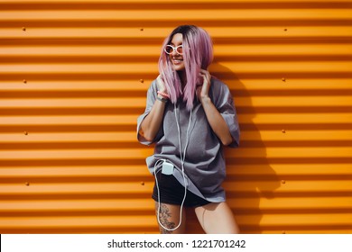 Beauty Woman Listening Music On Headphones, Outdoor Hipster Portrait, Purple Hair, Fashion Model, Color, Sunglasses, Smartphone, Tattoo, Sunglasses, Orange Wall, Happy Face, Smile, Hipster Style