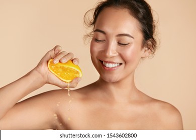 Beauty. Woman With Radiant Face Skin Squeezing Orange In Hand Portrait. Beautiful Smiling Asian Girl Model With Natural Makeup, Glowing Facial Skin And Citrus Fruit. Vitamin C Cosmetics Concept