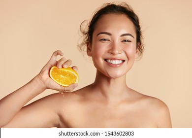 Beauty. Woman With Radiant Face Skin Squeezing Orange In Hand Portrait. Beautiful Smiling Asian Girl Model With Natural Makeup, Glowing Facial Skin And Citrus Fruit. Vitamin C Cosmetics Concept