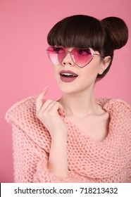Beauty wow. Fashion surprise teen girl model. Brunette in heart sunglasses with matte lips and bun hairstyle in knitted sweater posing over studio pink background.