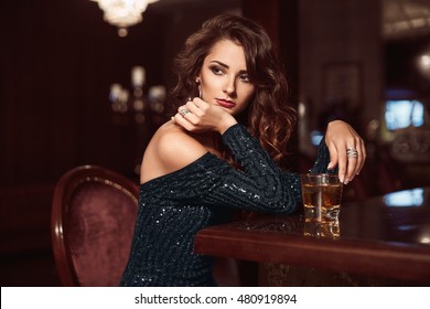 Beauty young brunette woman sitting at the bar with glass of whiskey in luxury interior