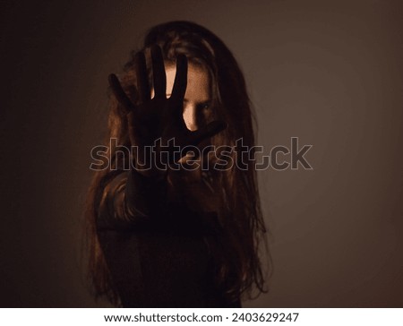 Beauty young witch with long dark hair conjures the hand covering the face on studio dark background in shadow light. Magic concept portrait. Focus on the palm. Toned color