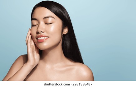 Beauty.Woman applying skincare product on her face. Asian girl with smooth perfect skin, touches her skin with pleased smile, enjoys facial cream rejuvenation effect, stands over blue background - Shutterstock ID 2281338487