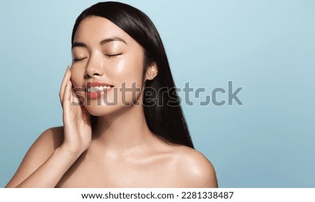 Beauty.Woman applying skincare product on her face. Asian girl with smooth perfect skin, touches her skin with pleased smile, enjoys facial cream rejuvenation effect, stands over blue background