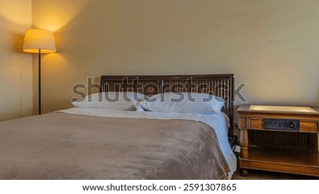 The bed is covered with white sheets and a beige terry-cloth bedspread. Two pillows at the wooden headboard. A floor lamp with a lampshade is lit. The bedside table is nearby. A fragment of a bedroom