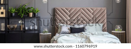 Bed with headboard between gold tables in grey bedroom interior with plants. Real photo