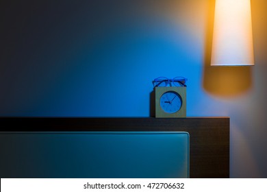 Bed Headboard, Over Head Lamp, Alarm Clock, And Reading Glasses At Night In Contrasting Cold Blur And Warm Orange Color Tones, In Simple And Minimalist Design With Room For Copy Space