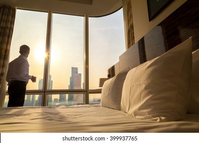 Bed maid-up with white pillows and bed sheets in cozy room. Young businessman with cup of coffee standing at window looking at city scenery on the background. Focus on cushion. Motivation concept