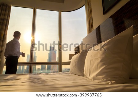 Bed maid-up with white pillows and bed sheets in cozy room. Young businessman with cup of coffee standing at window looking at city scenery on the background. Focus on cushion. Motivation concept