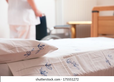 Bed At Nursing Home With Nurse And Wheelchair On Background.