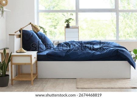 Bed with stylish blue bedding, commode, bedside table and houseplants near window in bedroom