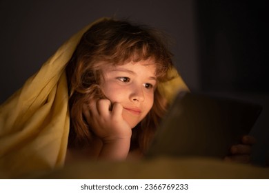 Bed time story. Kid playing on a digital tablet. Kid using tablet for gaming and online learning while lying on the bed, screen light reflex on her face. Child play tablet under blanket at night. - Powered by Shutterstock