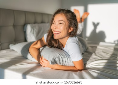 Bed Woman Waking Up Early Morning Happy Enjoying Sun On Comfortable Mattress And Pillow. Asian Girl Relaxing In Bedroom Smiling. Natural Beauty After Healthy Good Night Sleep Sleeping In On Weekend.