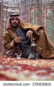 Bedouin Man Wearing Traditional Clothes Praying With A Tasbih While Drinking Tea On A Carpet In The Saudi Desert During The Night, Saudi Arabia