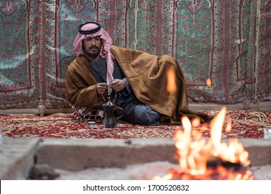 Bedouin Man Wearing Traditional Clothes Praying With A Tasbih While Drinking Tea On A Carpet In Front Of A Fire In The Saudi Desert, Saudi Arabia