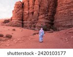 A bedouin tribesman walks on a desert path in traditional Middle Eastern clothing towards an opening in a distant canyon, with rich red sand and water eroded sandstone during the hot summer