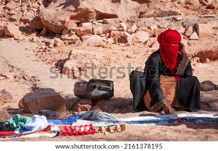Bedouin woman with covered face sits on sand against rocks in desert selling souvenirs to tourists in shadow. Bedouin village in Sahara on hot sunny day in summer