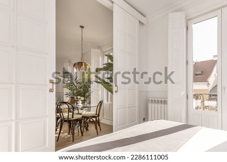 Bedroom with a balcony with glass and white wood doors and sliding wooden doors leading to a dining room with a circular glass table