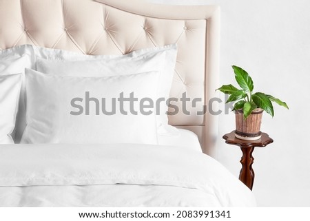 Bedroom with bed, white bedding, and bedside table with indoor flower in a pot. White pillows, duvet and duvet case on bed with beige headboard.  Bed with clean white pillows and bed sheets.