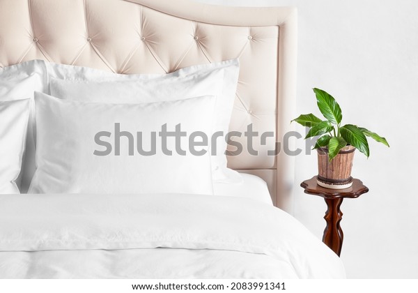 Bedroom with bed, white bedding, and bedside\
table with indoor flower in a pot. White pillows, duvet and duvet\
case on bed with beige headboard.  Bed with clean white pillows and\
bed sheets.