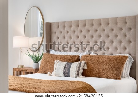 A bedroom detail shot with a cushioned headboard, a gold mirror on the wall, and a plant and lamp on the nightstand.