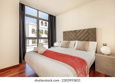 A bedroom with a double bed with a large window overlooking the street and a headboard upholstered in gray velvet