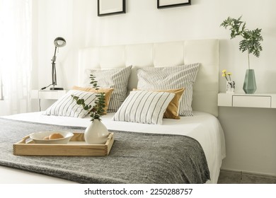 Bedroom with a double bed with a white leather upholstered headboard with cushions of various types and a wooden tray with a vase and rolls in a white bowl