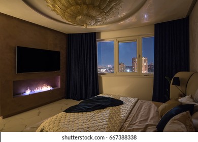 Bedroom In A Modern Style With A Big Fancy Golden Chandelier On The Ceiling. There Is A Bed With Pillows And A Coverlet, Burning Fireplace, TV On The Wall, Windows With Curtains. Indoors. Horizontal.