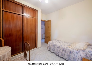 Bedroom with small bed with ugly patterned bedspread and large built-in wardrobe with sliding doors made of dark varnished wood