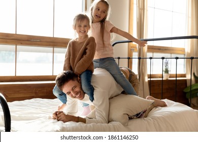 In Bedroom Two Little Cheery Daughters Play With Dad, Saddled Him Seated On His Back. Laughing Father Giving Piggy Back Ride To Children Enjoy Funny Activity In Morning At Home. Family Games Concept