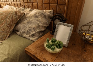 A bedside table holds a clock and an empty frame beside a bowl of succulent plants. The setting shows a soft bed with pillows and a warm wooden table, suggesting a relaxed space.