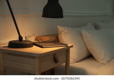 Bedside table with lamp, notebook, glasses and glass of water near bed indoors