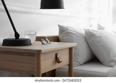 Bedside table with lamp, notebook, glasses and glass of water near bed indoors, closeup