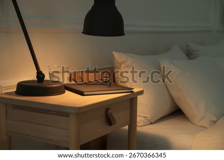 Bedside table with lamp, notebook, glasses and glass of water near bed indoors