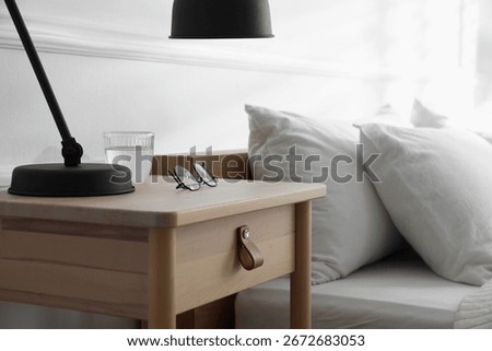 Bedside table with lamp, notebook, glasses and glass of water near bed indoors, closeup