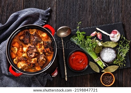 Beef Bourguignon, Beef Burgundy, stew with beef, bacon, carrots, onions and mushrooms slow cooked in rich red wine sauce in pot on wooden table with ingredients, horizontal view from above, flat lay